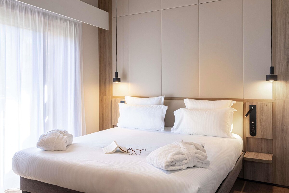 Une chambre d’hôtel avec un grand lit, deux tables de chevet, une tête de lit en bois et une fenêtre ornée de rideaux légers.