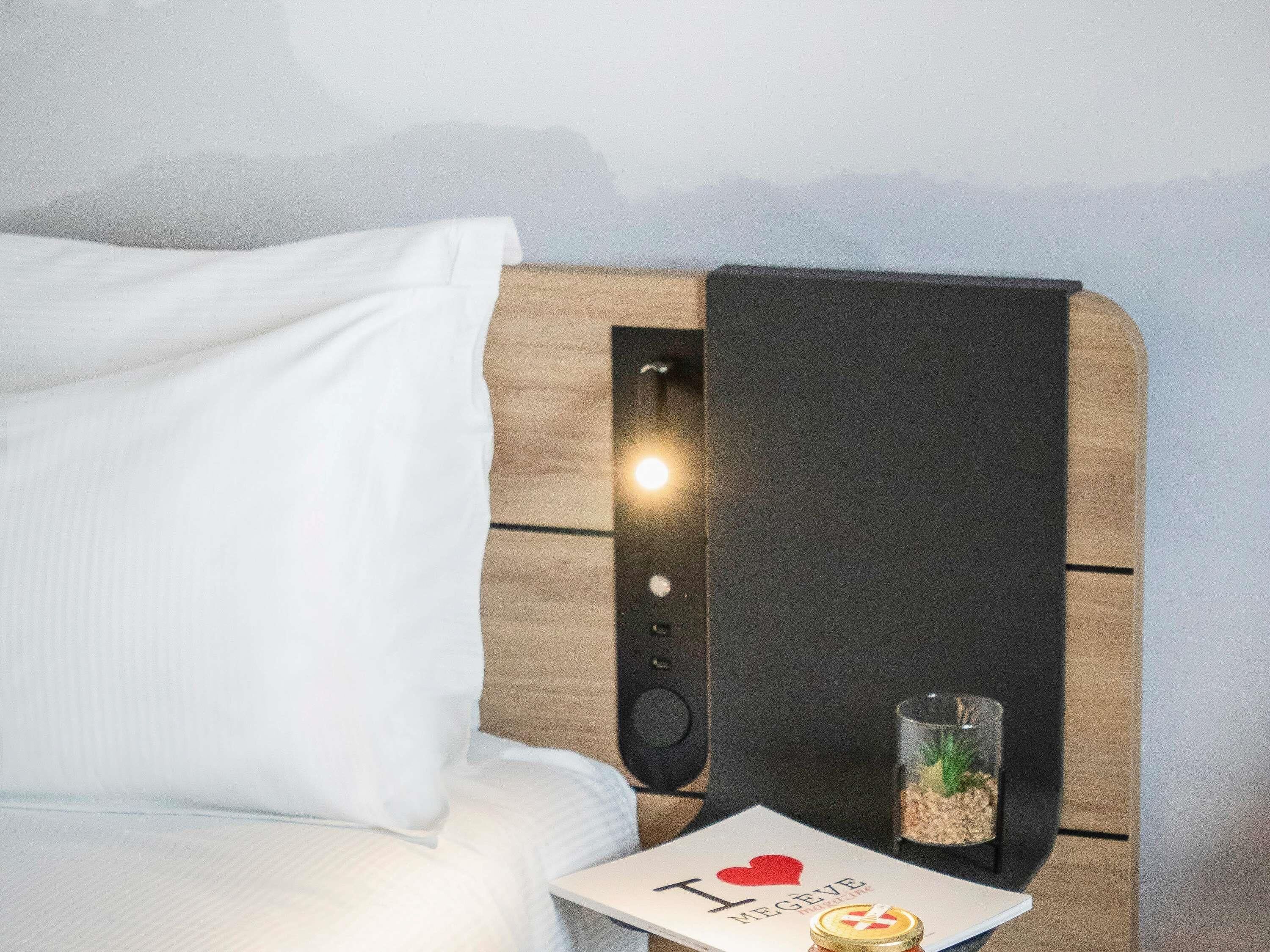 A neatly made bed with white linens, a bedside table with a small decorative item, and a wall-mounted light fixture.