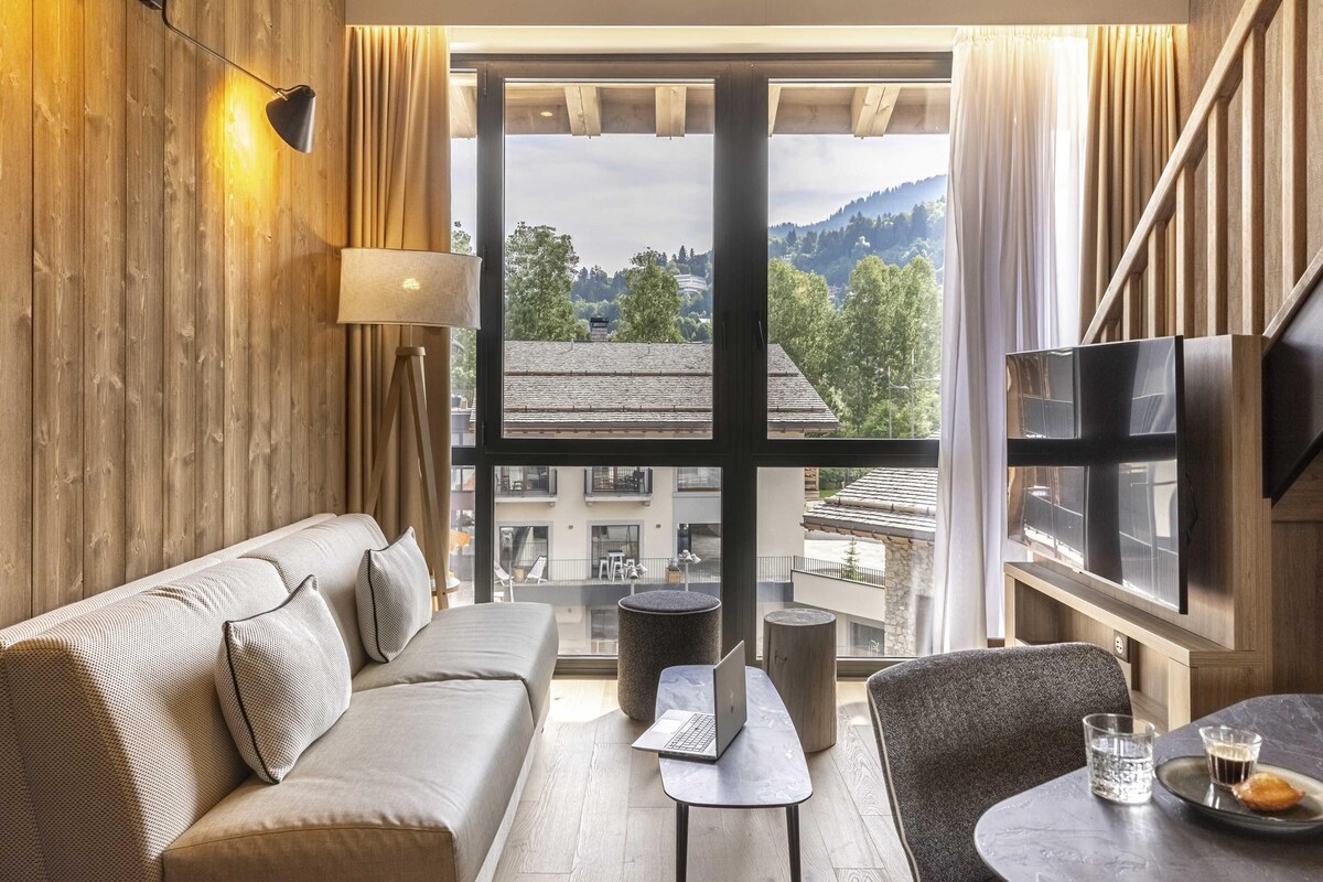 Une chambre d’hôtel moderne équipée d’un canapé, d’une petite table avec un ordinateur portable, d’une chaise, d’une télévision et d’une grande fenêtre donnant sur la ville.
