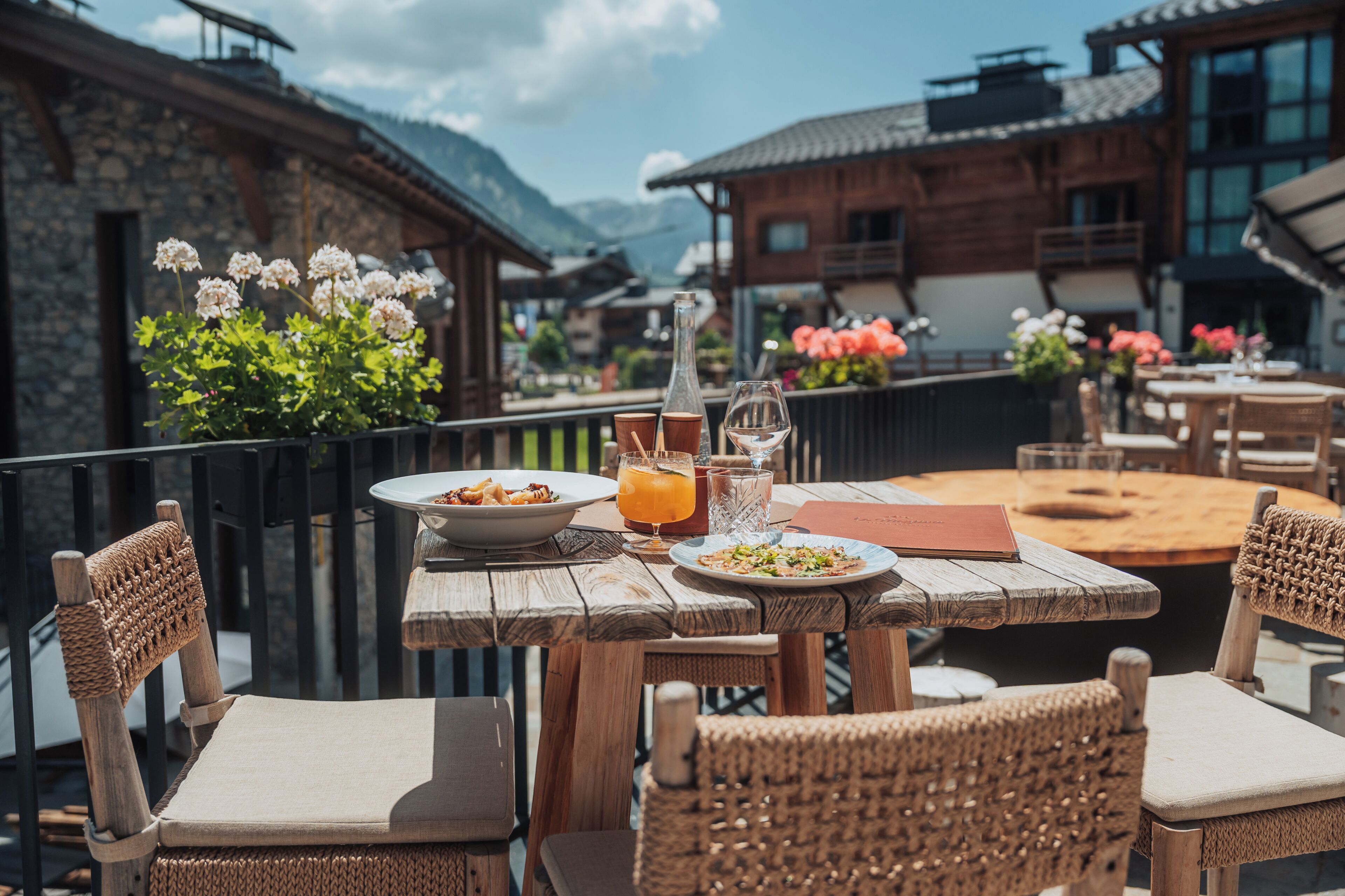 An outdoor dining area with wooden tables and wicker chairs, overlooking a mountainous landscape.
