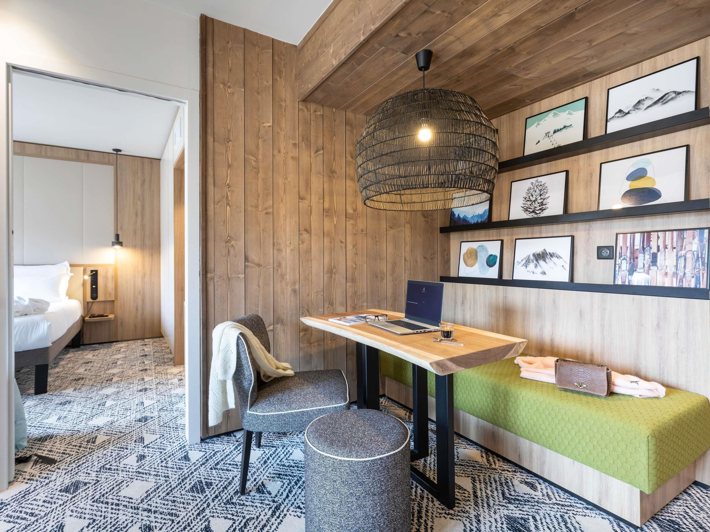 Une chambre d’hôtel moderne avec un mur en bois, une table ronde sur laquelle se trouve un ordinateur portable, un canapé vert et un grand luminaire suspendu.