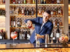Un barman prépare un cocktail dans un bar, avec des étagères remplies de bouteilles d’alcool en arrière-plan.