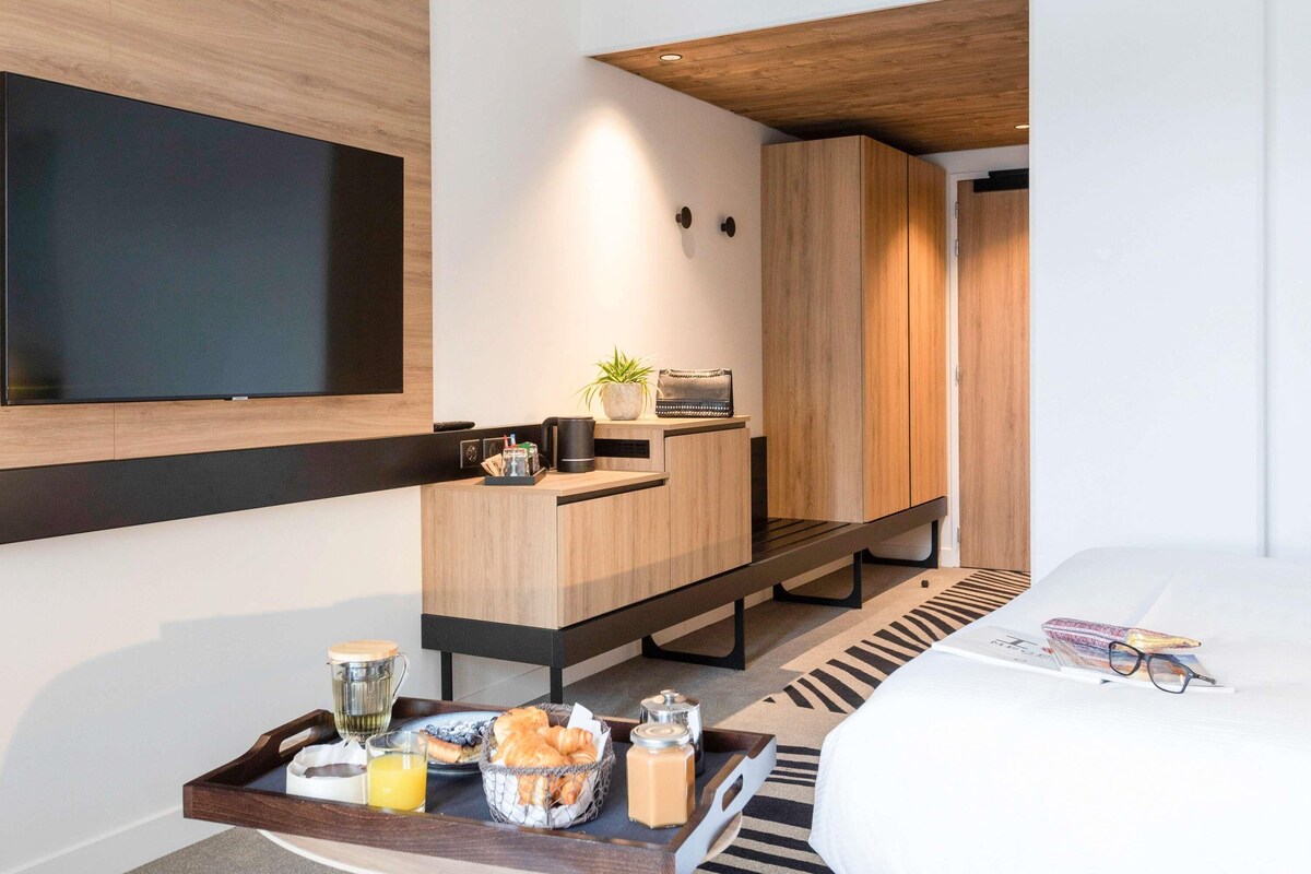 Une chambre d’hôtel moderne dotée d’une télévision à écran plat, de meubles en bois, d’un plateau de petit-déjeuner avec des croissants et d’un lit agrémenté d’un magazine et de lunettes.