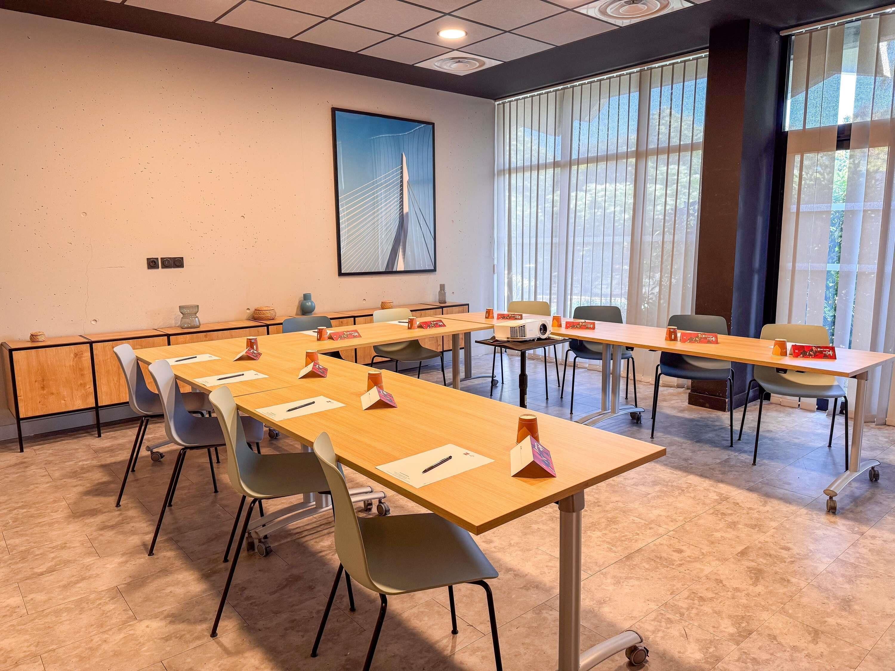 A conference room with long tables, chairs, a projector, and a large window with blinds.