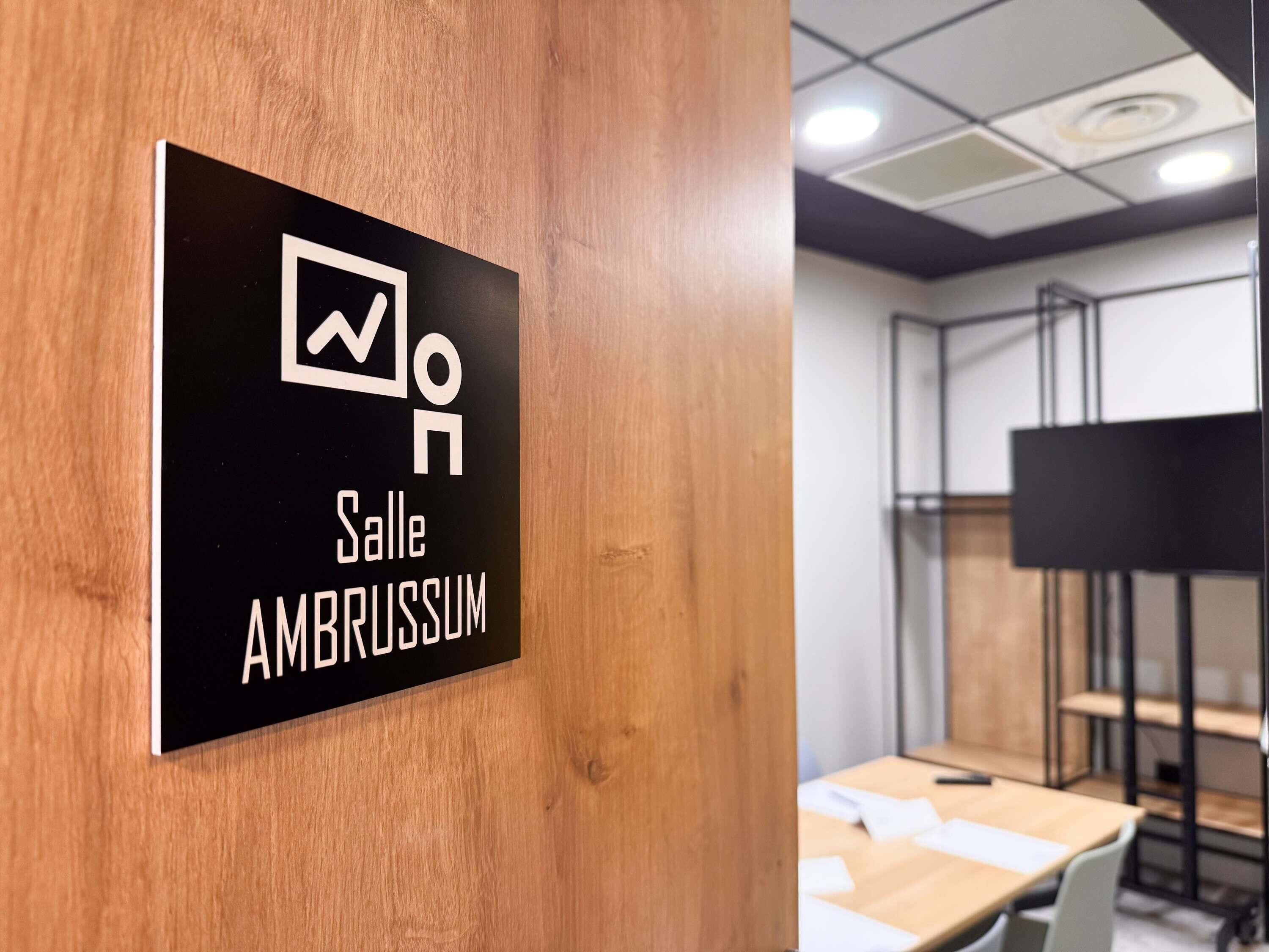 A sign indicating 'Salle Ambussum' in a room with wooden paneling and a white ceiling.