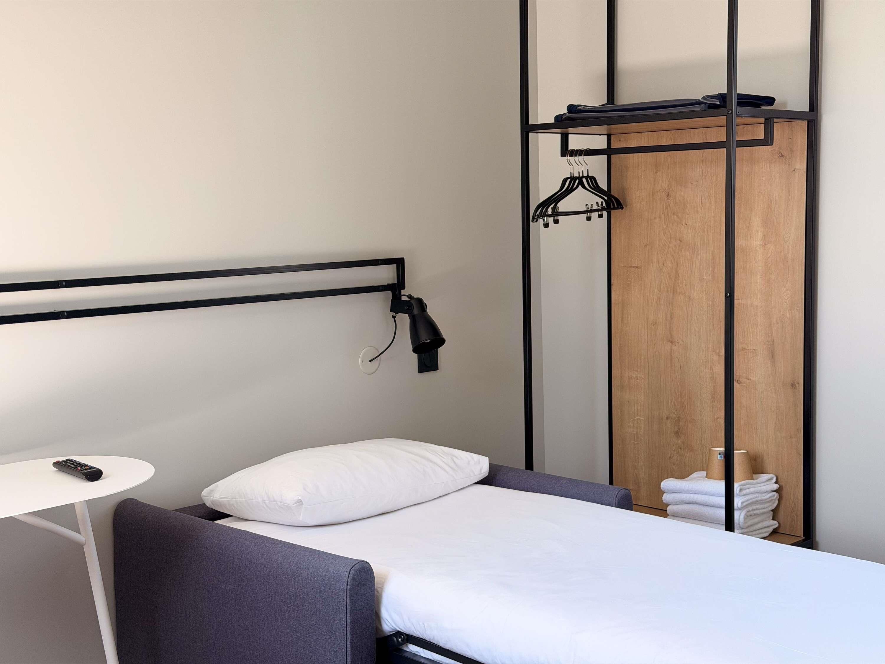 A modern hotel room with a bed, a small table, a wall-mounted lamp, and a wooden wardrobe with a hanging rack.