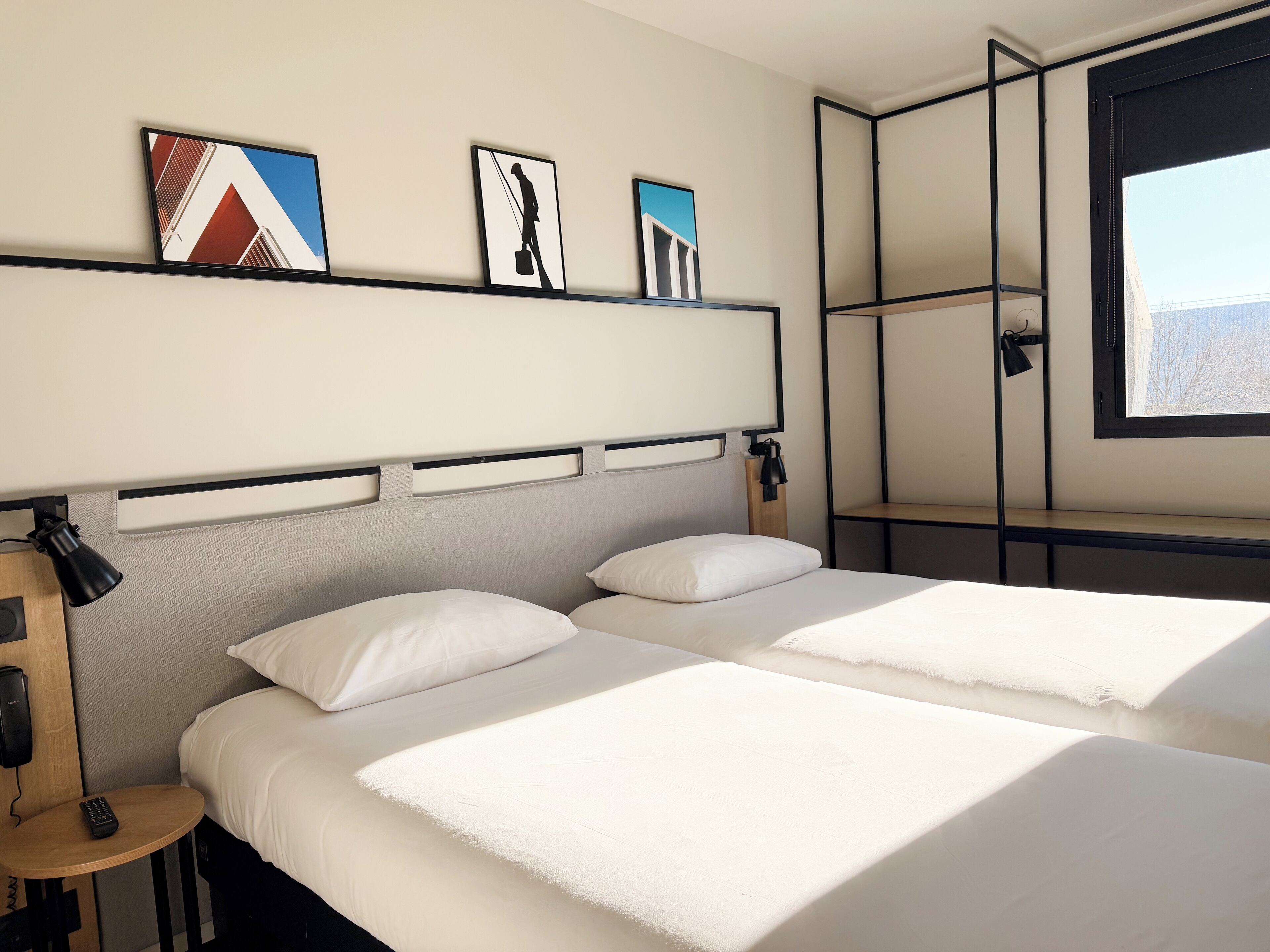 A hotel room with two beds, a bedside table with a lamp, a wall-mounted shelf with artwork, and a window with a view.