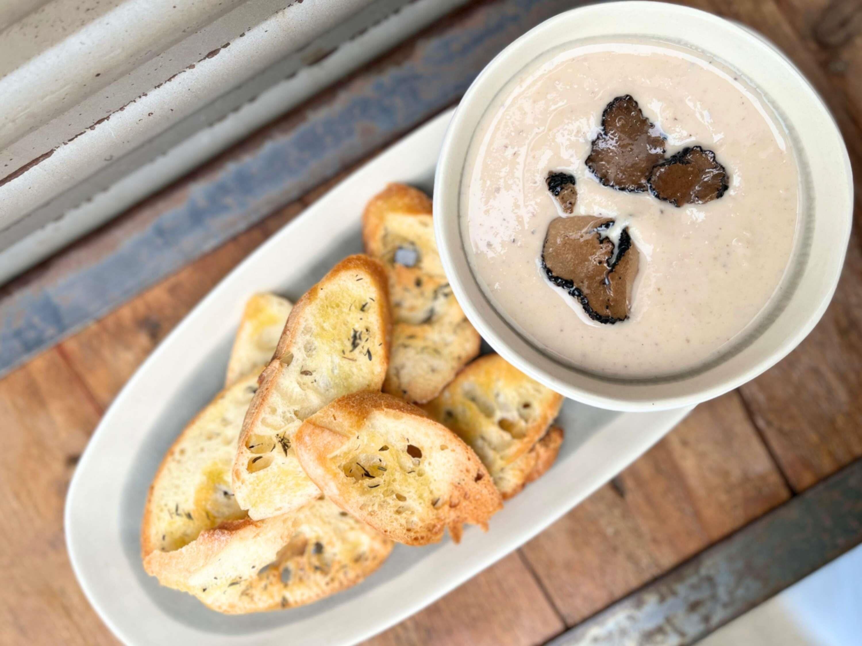 A bowl of soup with a creamy texture and a few dark pieces on top, accompanied by several slices of garlic bread on a white plate.
