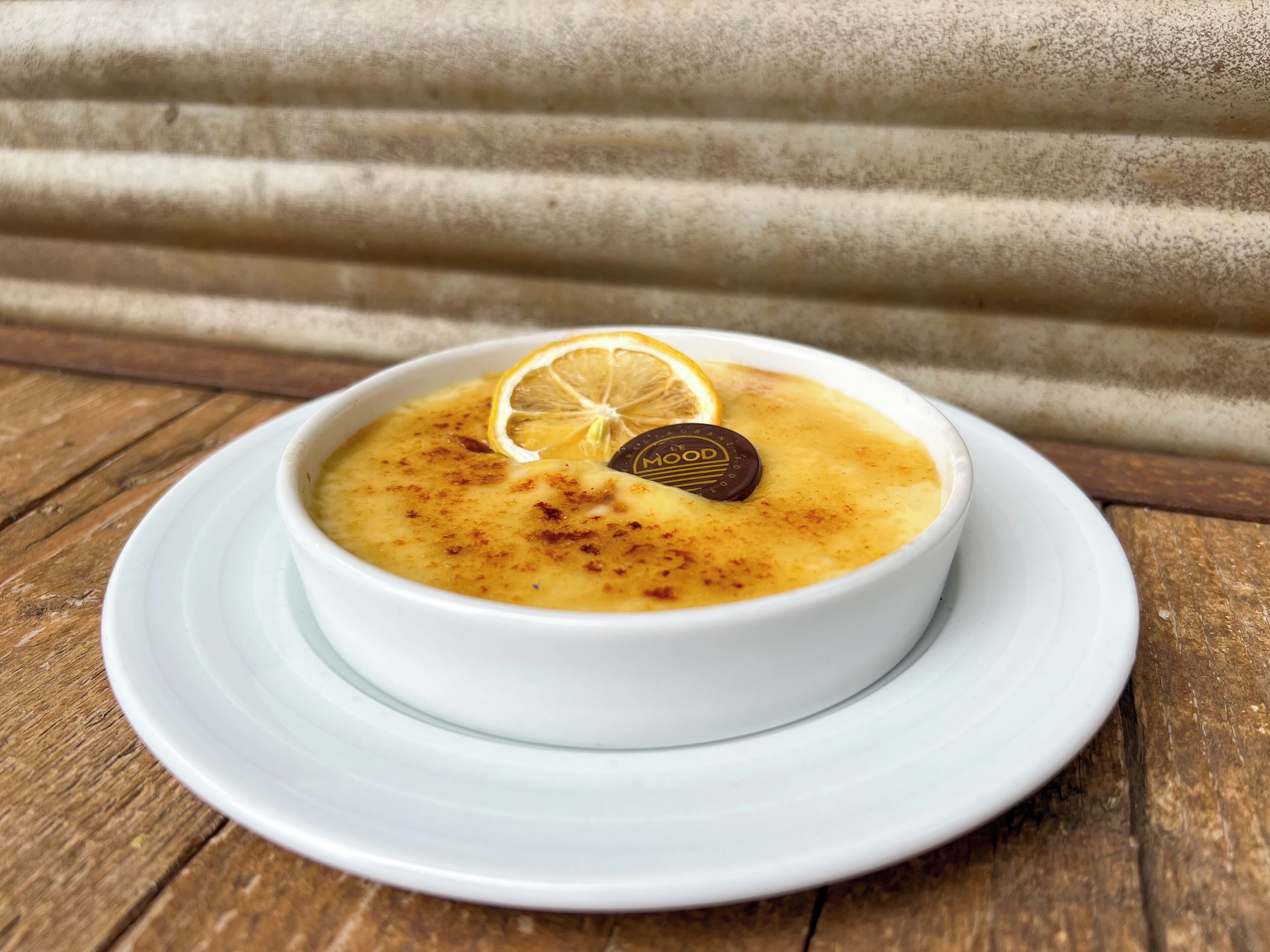 Uma tigela de sopa sobre um prato branco, com uma fatia de limão e um pedaço de chocolate por cima, colocada sobre uma mesa de madeira.