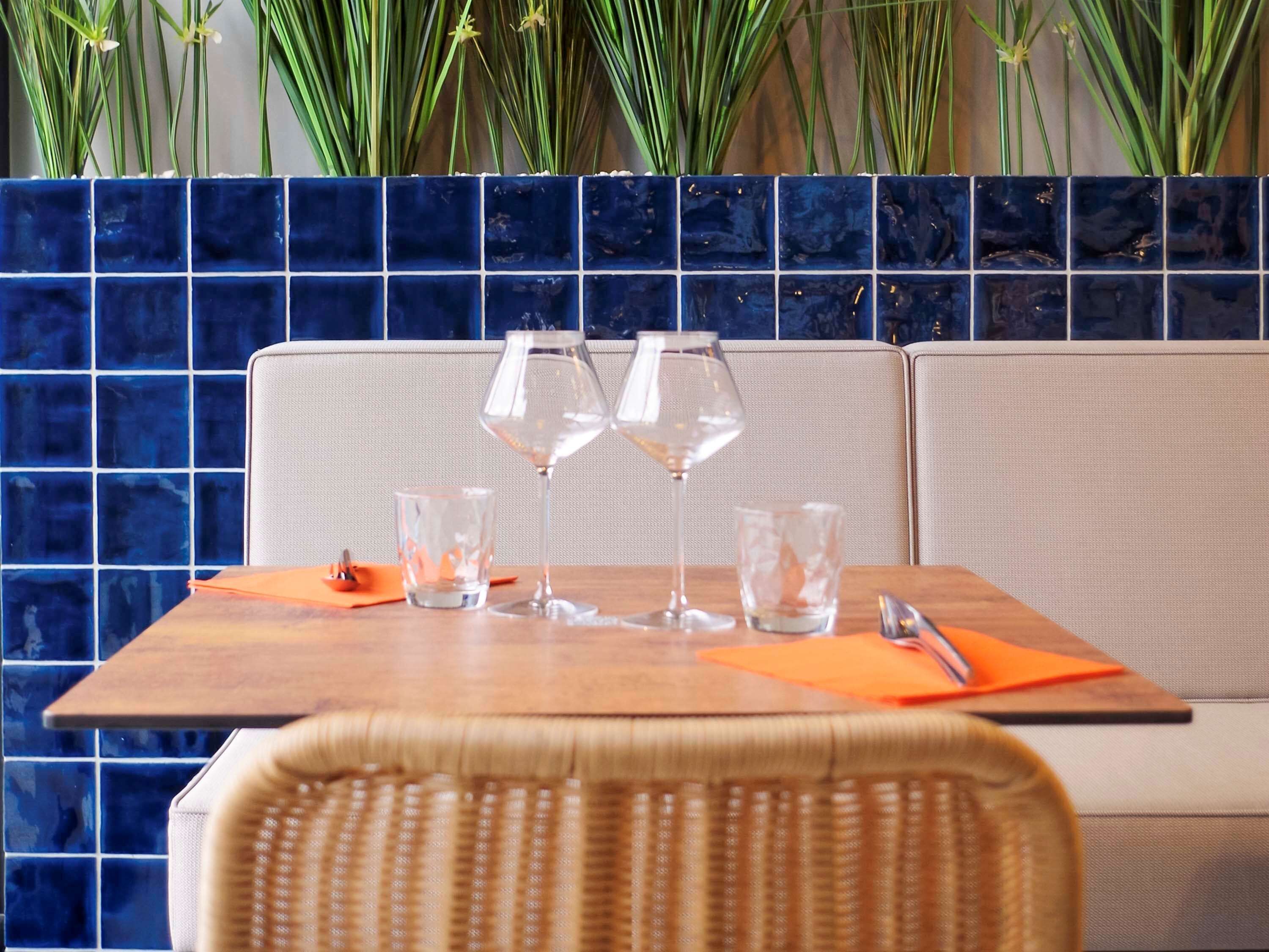 A dining area with a table set for two, featuring a woven chair, a blue tiled backsplash, and a plant with green leaves and yellow flowers.
