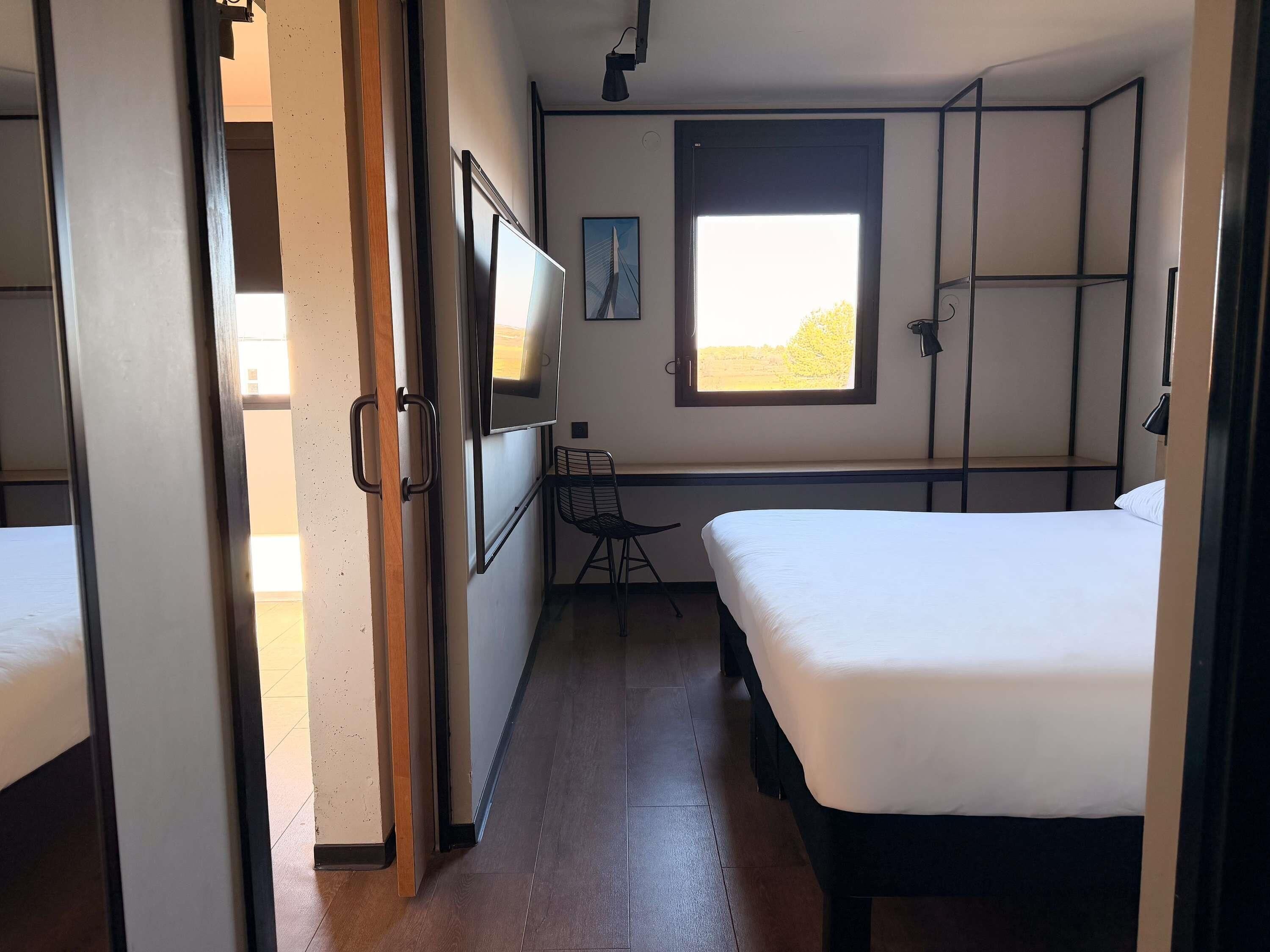 Una habitación con dos camas, un escritorio, una silla, un ventanal con vistas y una puerta que da a otra habitación.
