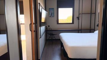 Una habitación con dos camas, un escritorio, una silla, un ventanal con vistas y una puerta que da a otra habitación.