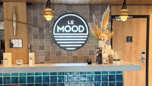 A hotel reception area with a blue tiled counter, a wooden reception desk, a neon sign reading "LE MOOD", and a modern light fixture. - Ibis Lunel Petite Camargue (Lunel)