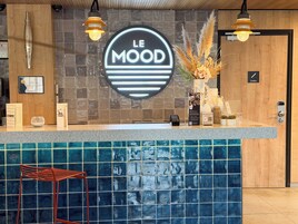 Zona de recepción de hotel con un mostrador azul de azulejos, un escritorio de recepción de madera, un letrero de neón que dice "LE MOOD" y una lámpara moderna.