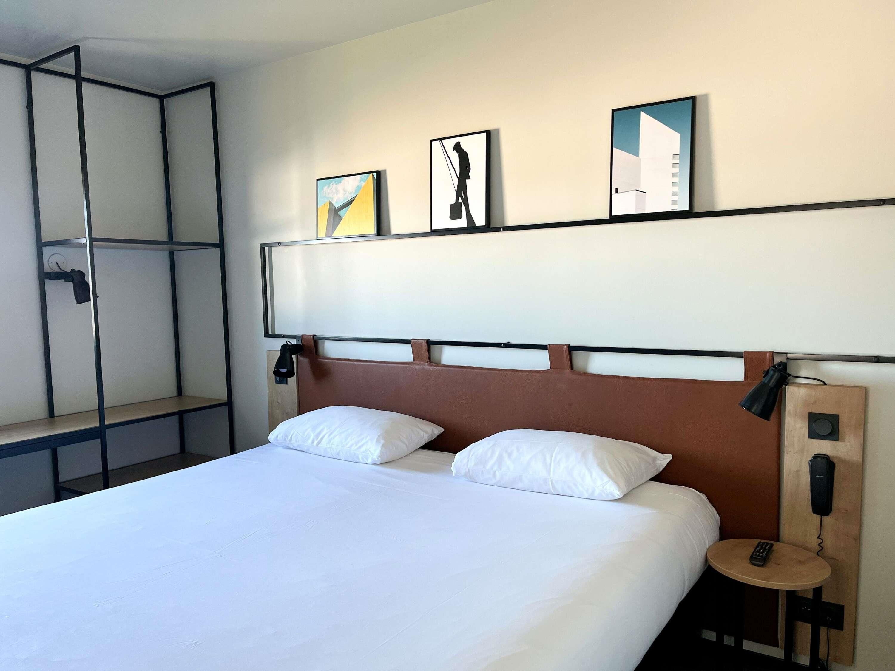 Une chambre d’hôtel comprenant un lit, deux oreillers, une table de chevet avec une télécommande, un téléphone fixé au mur et trois œuvres d’art encadrées au-dessus du lit.