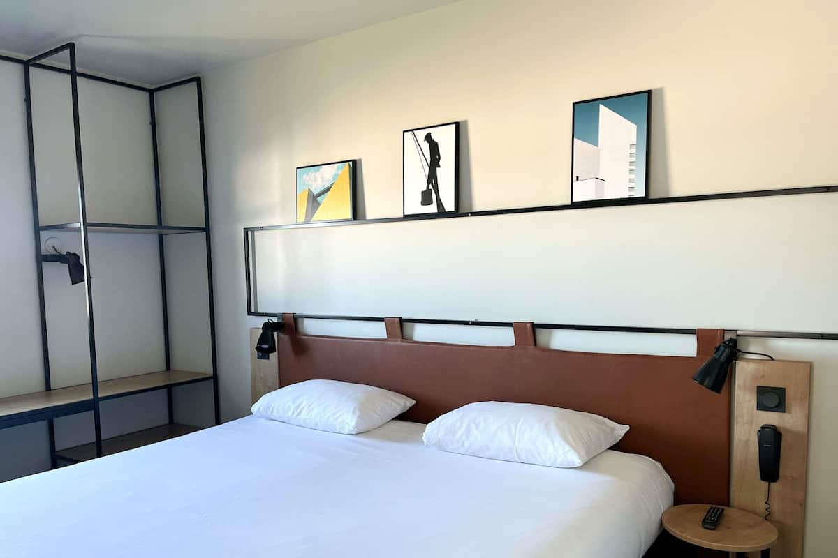 A hotel room with a bed, two pillows, a bedside table with a remote control, a wall-mounted phone, and three framed artworks above the bed.