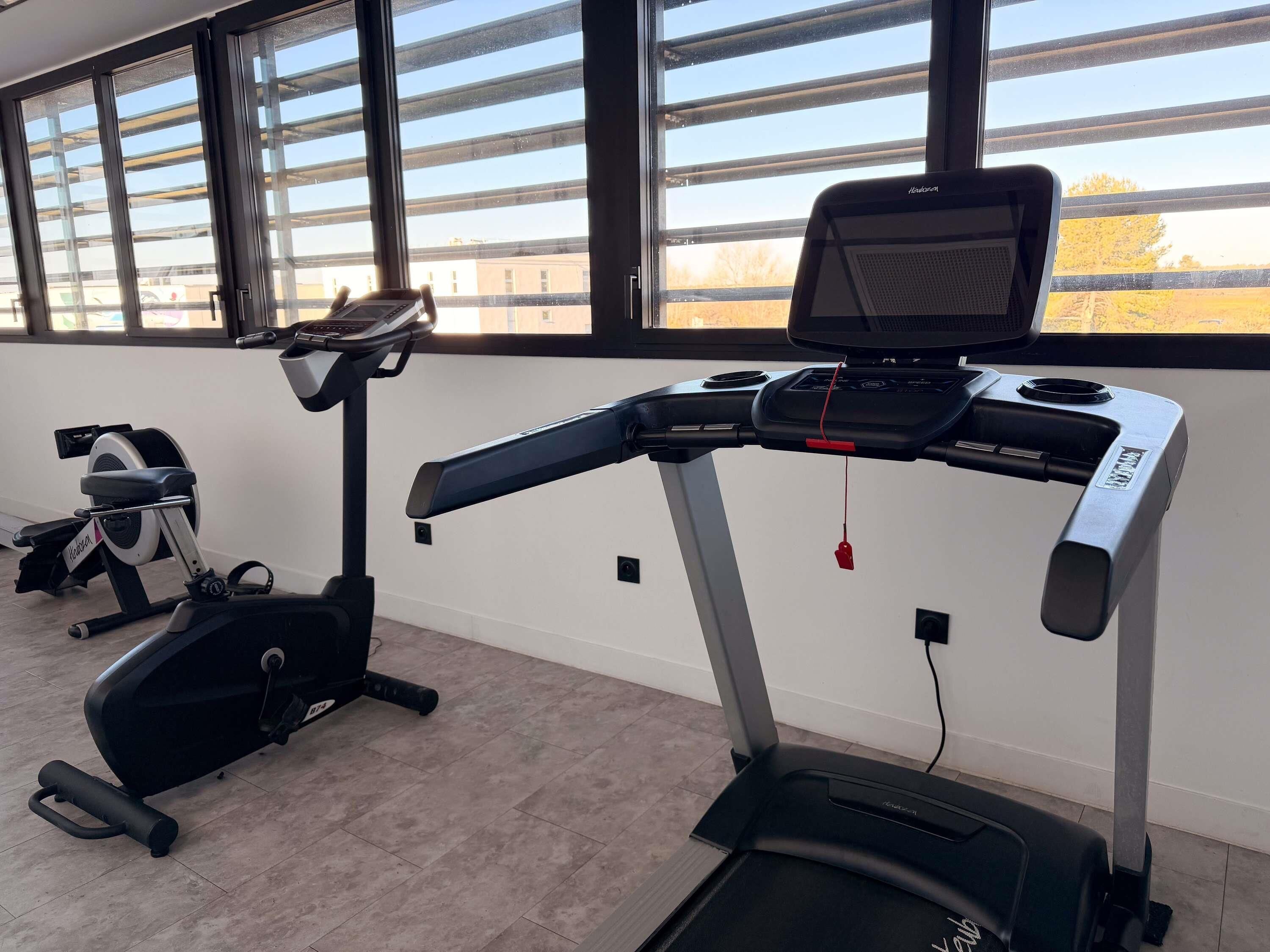 Un gimnasio con cintas de correr y equipo de ejercicio, amplias ventanas con vistas a edificios, y un interior moderno y limpio.