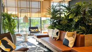 A modern lounge area with a sofa, chairs, and a round table, featuring a geometric patterned rug and large windows with blinds. - Ibis Lunel Petite Camargue (Lunel)