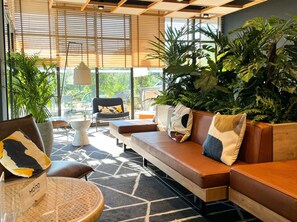 Een moderne zithoek met een bank, stoelen en een ronde tafel, met een vloerkleed met geometrische patronen en grote ramen met luxaflex.