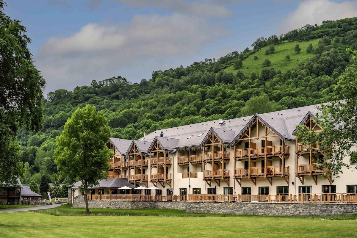 Un hotel de varios pisos con balcones de madera y fachada de piedra, con colinas verdes exuberantes al fondo.