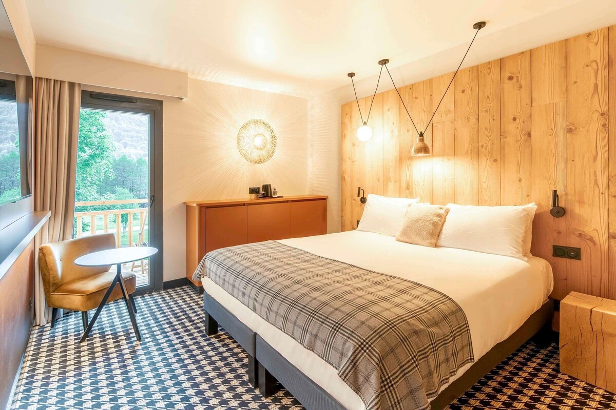 Een hotelkamer met een bed, een klein tafeltje, een stoel, een houten hoofdbord en uitzicht op het landschap.