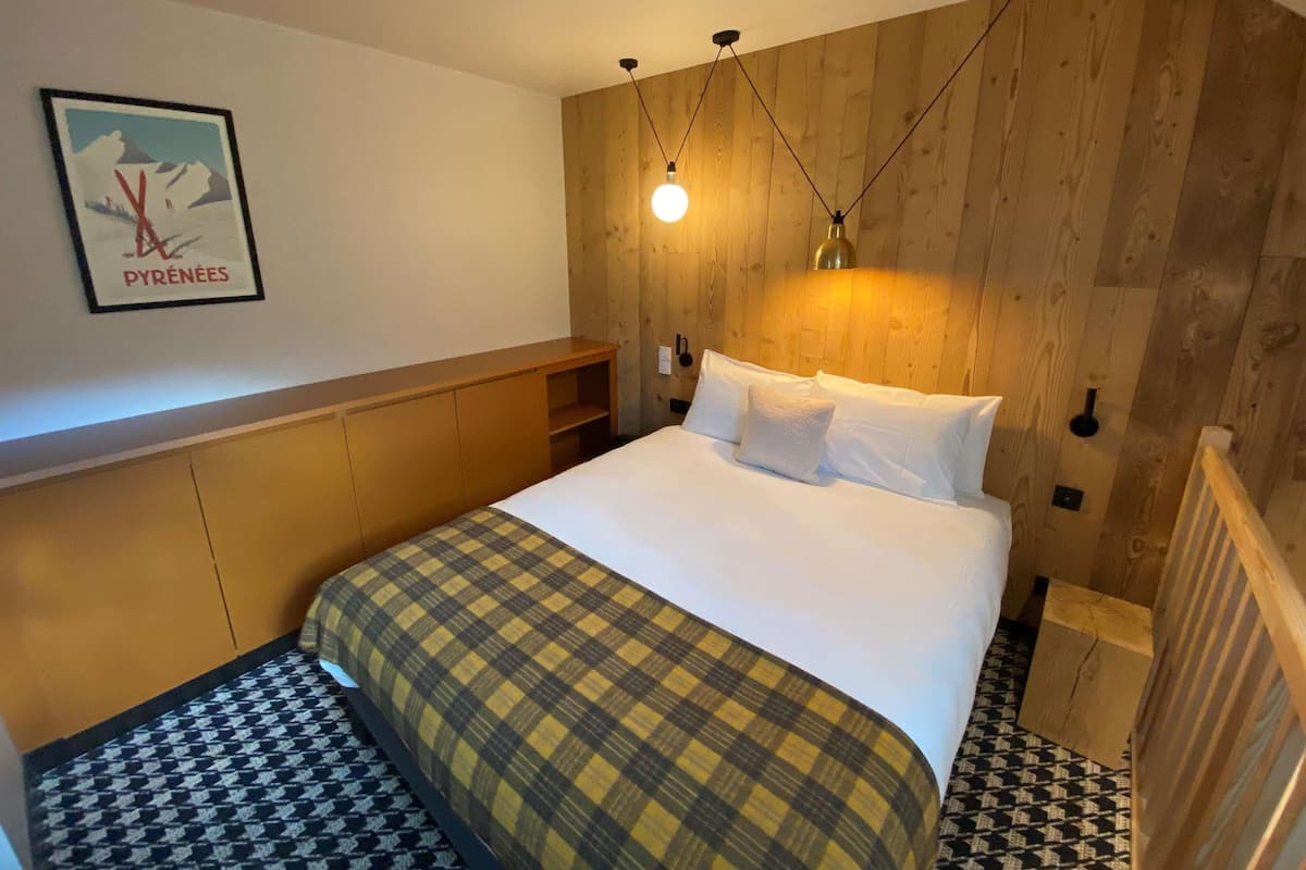 Une chambre à coucher avec une tête de lit en bois, un lit recouvert de draps blancs, une couverture à carreaux et un luminaire fixé au mur.