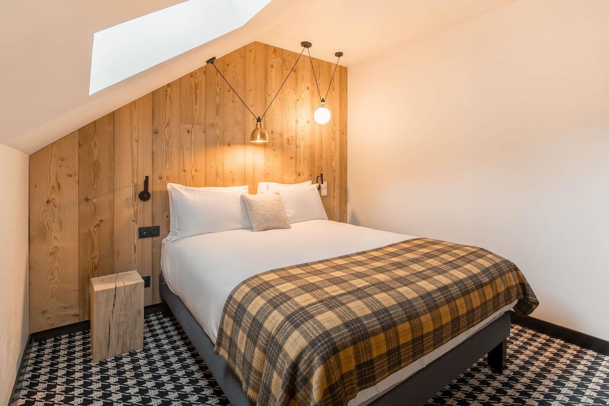Une chambre avec un plafond en pente, des murs lambrissés, un lit avec une literie blanche, une couverture à carreaux et un sol à motifs.