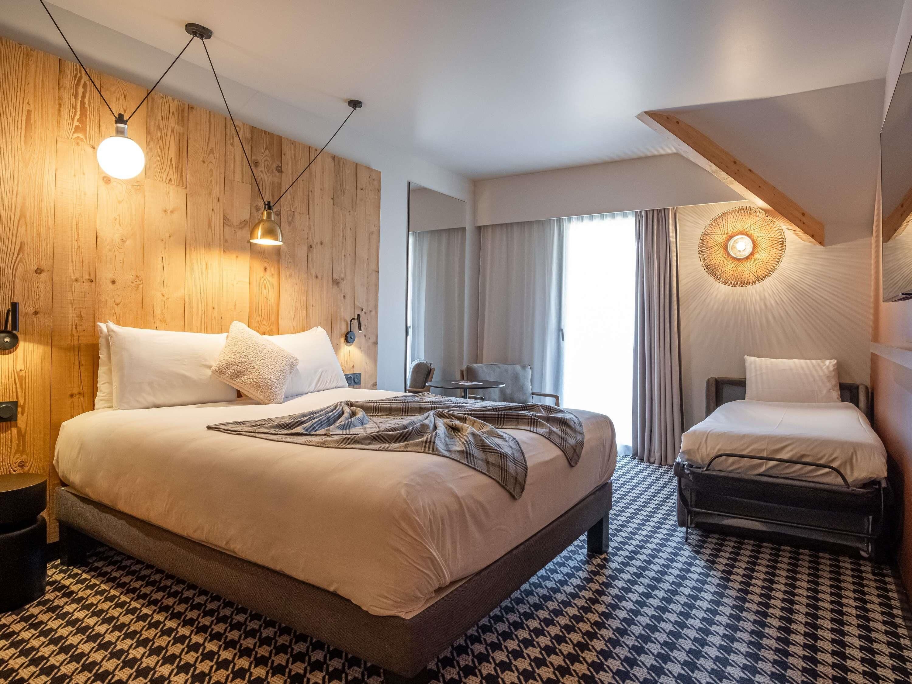 Une chambre d’hôtel avec un grand lit, un plus petit lit, un bureau et une chaise. Il y a une fenêtre avec des rideaux, une lampe fixée au mur et un luminaire au plafond.