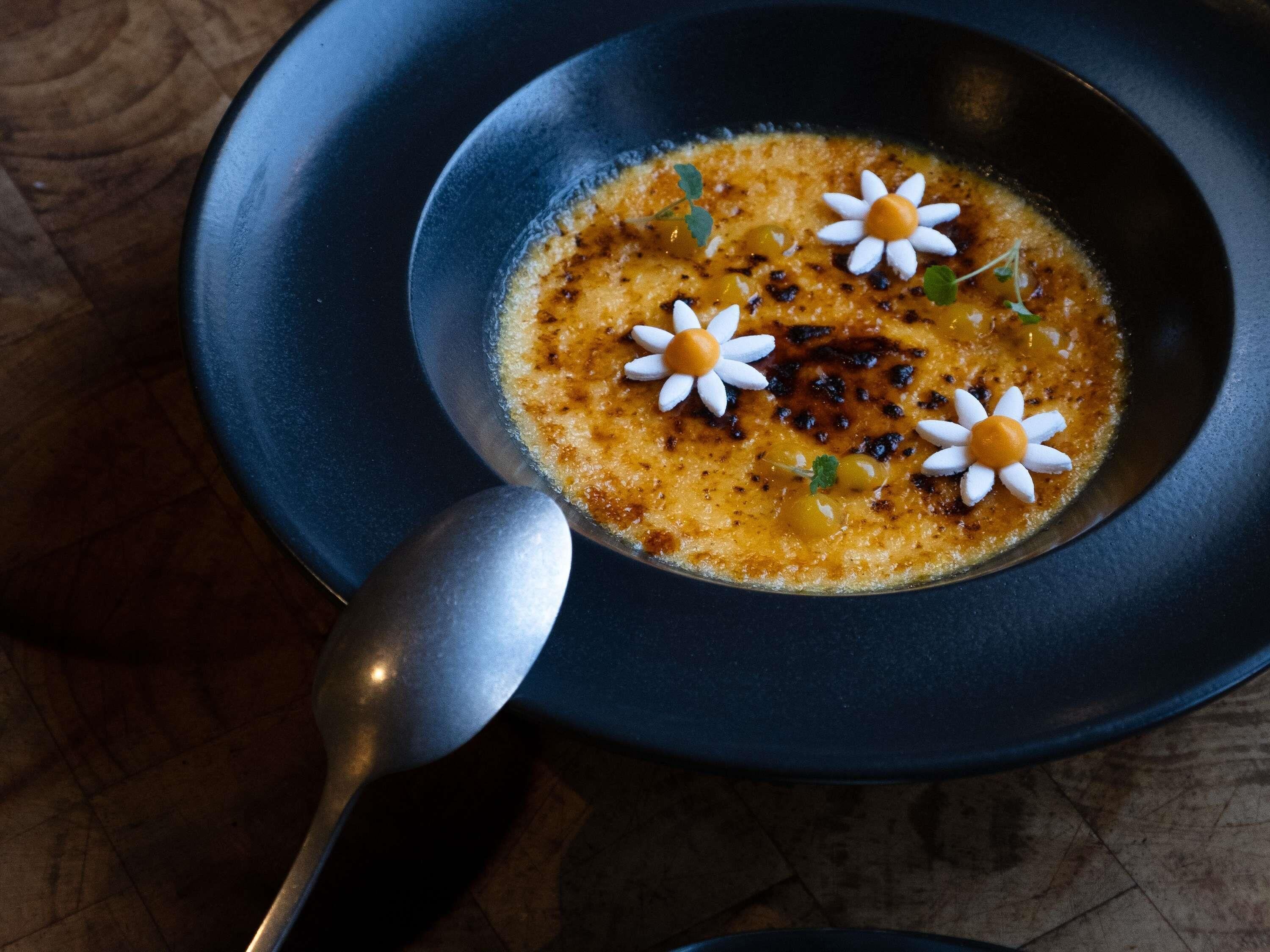 Un tazón de sopa con una cuchara, adornado con flores, sobre un plato oscuro.