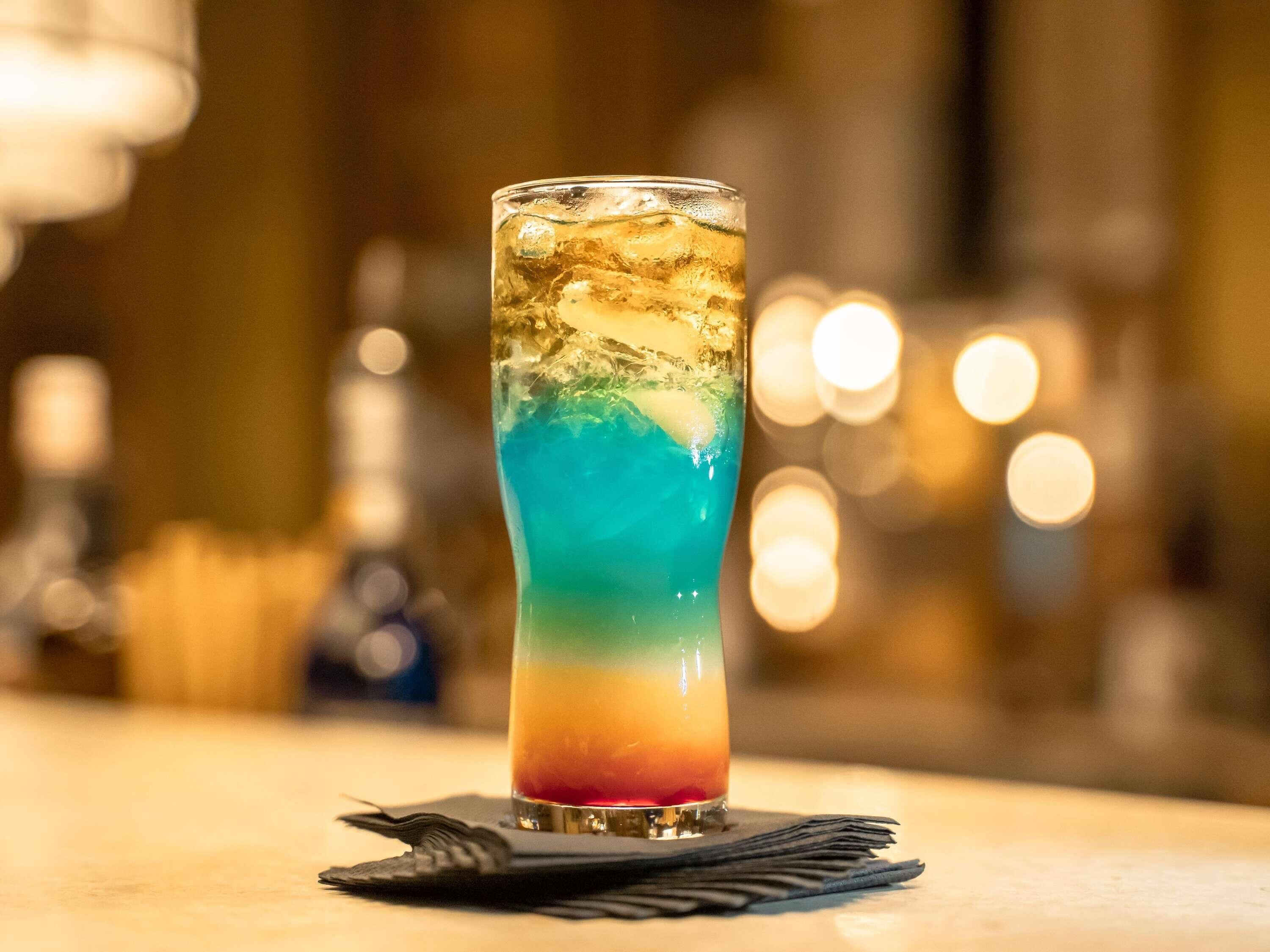 A tall glass with a layered drink, ice cubes, and a cocktail napkin on a bar counter.