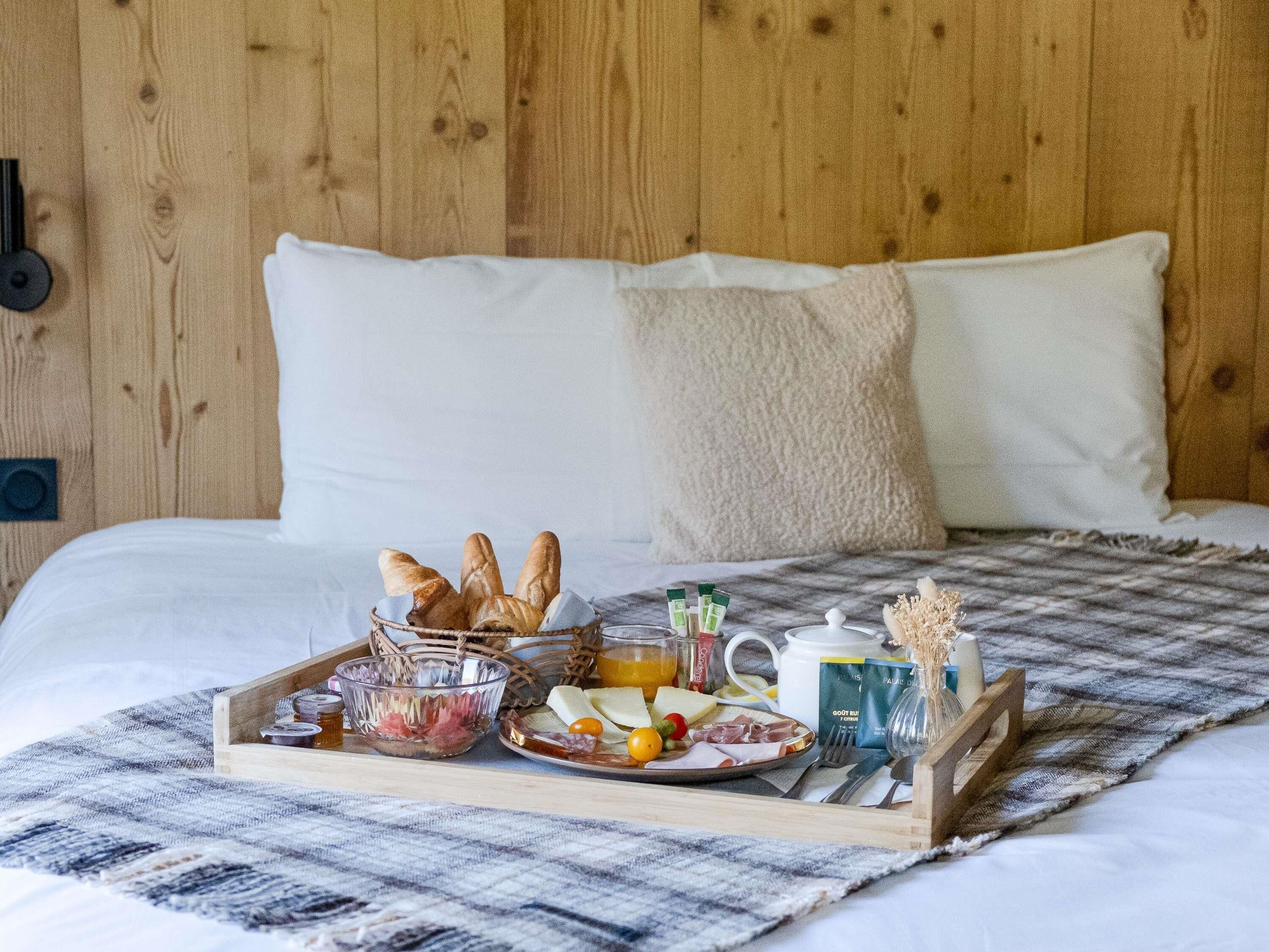Una bandeja de desayuno bien dispuesta sobre una cama con una manta escocesa, que incluye pan, queso, fruta y bebidas.