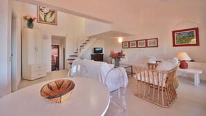 A modern living room with a white sofa, a wicker chair, a dining table with a woven bowl, a staircase, and artwork on the walls.