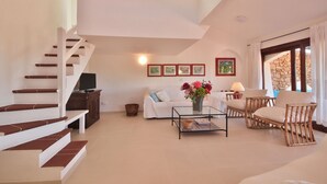 A spacious living area with a staircase, white sofa, wicker chairs, a coffee table, and artwork on the wall.