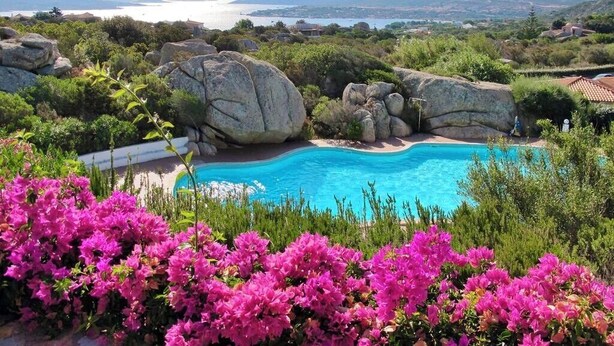 A pool surrounded by large rocks and pink flowers, with a view of the sea and distant hills.