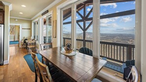 A dining area with a wooden table and chairs, a view of the landscape through large windows, and a balcony with a railing. - The Treetop Terrace, VIEWS, DECKS, SLEEPS 13 (West Jefferson)