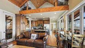 A spacious living area with a leather sofa, wooden dining table, and a kitchen in the background. - The Treetop Terrace, VIEWS, DECKS, SLEEPS 13 (West Jefferson)