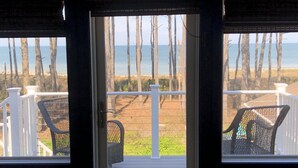 A view from a room with a door leading to a balcony overlooking a beach with tall trees and the ocean.