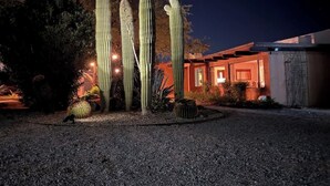 Vista nocturna de un edificio con cactus altos al frente, un camino de grava y un exterior con luz cálida.