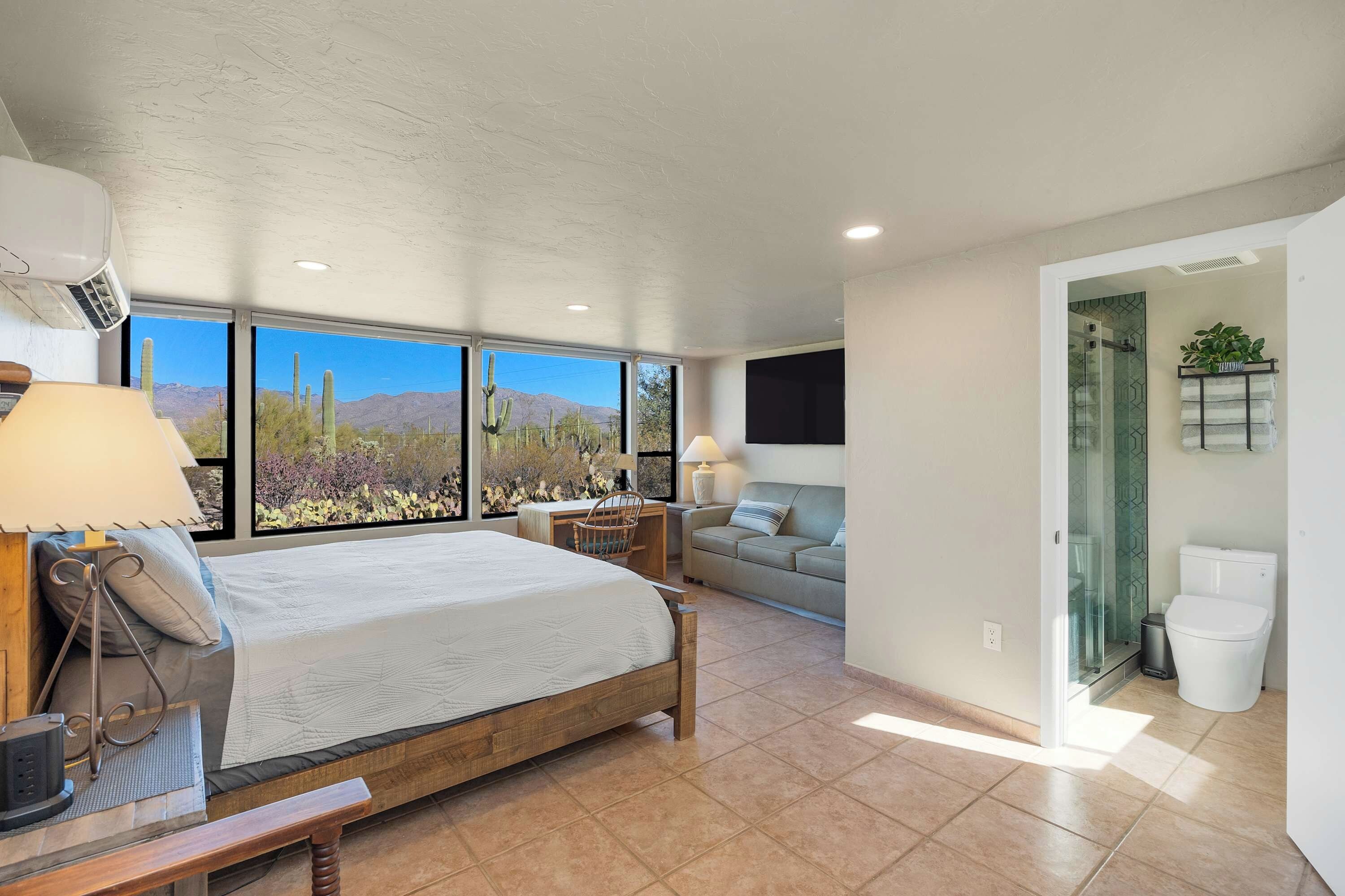 A bedroom with a large bed, a desk, a chair, a sofa, and a shower area.