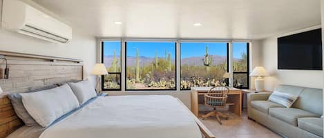 A bedroom with a large bed, a desk, a chair, and a sofa. There are windows with a view of cacti and mountains.
