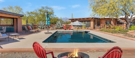 A backyard with a swimming pool, red chairs, a fire pit, and a covered patio area.