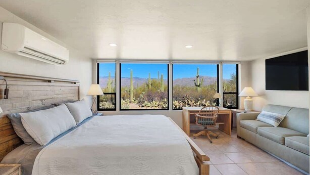 Un dormitorio con una cama grande, un escritorio, una silla y un sofá. Hay ventanas amplias con vistas a cactus y montañas.