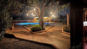 Un espacio exterior nocturno con piscina, rodeado de árboles e iluminado por luces colgantes.