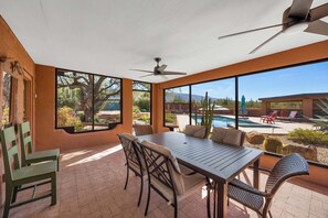 A sunlit patio with a table and chairs, a ceiling fan, and a view of a swimming pool and desert landscape. - Saguaro Retreat-Salt Pool, Spa, Mountain Views (Tucson)