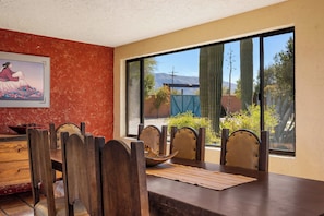 A dining area with a wooden table and chairs, a painting on the wall, and a view of cacti and a mountain in the background. - Saguaro Retreat-Salt Pool, Spa, Mountain Views (Tucson)