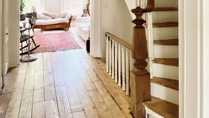 Un pasillo amplio con una escalera de madera que sube a un segundo piso. Se ve una mecedora y un ventilador al frente.