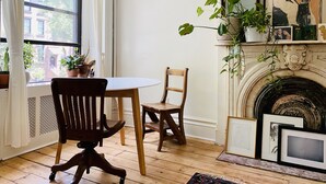Una habitación con una mesa y sillas de madera, una chimenea con obras de arte enmarcadas, y una ventana con cortinas.