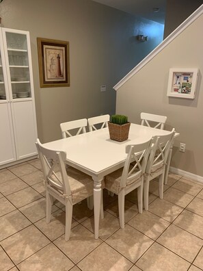 Un comedor con una mesa y sillas blancas, una cesta de mimbre con una planta, un cuadro en la pared y un mueble con puertas de cristal.