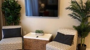 A living room with two patterned armchairs, a small coffee table, a flat-screen TV mounted on the wall, and two potted plants.