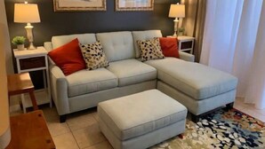A living room with a light-colored sectional sofa, two side tables with lamps, a small wooden stool, and a patterned rug.